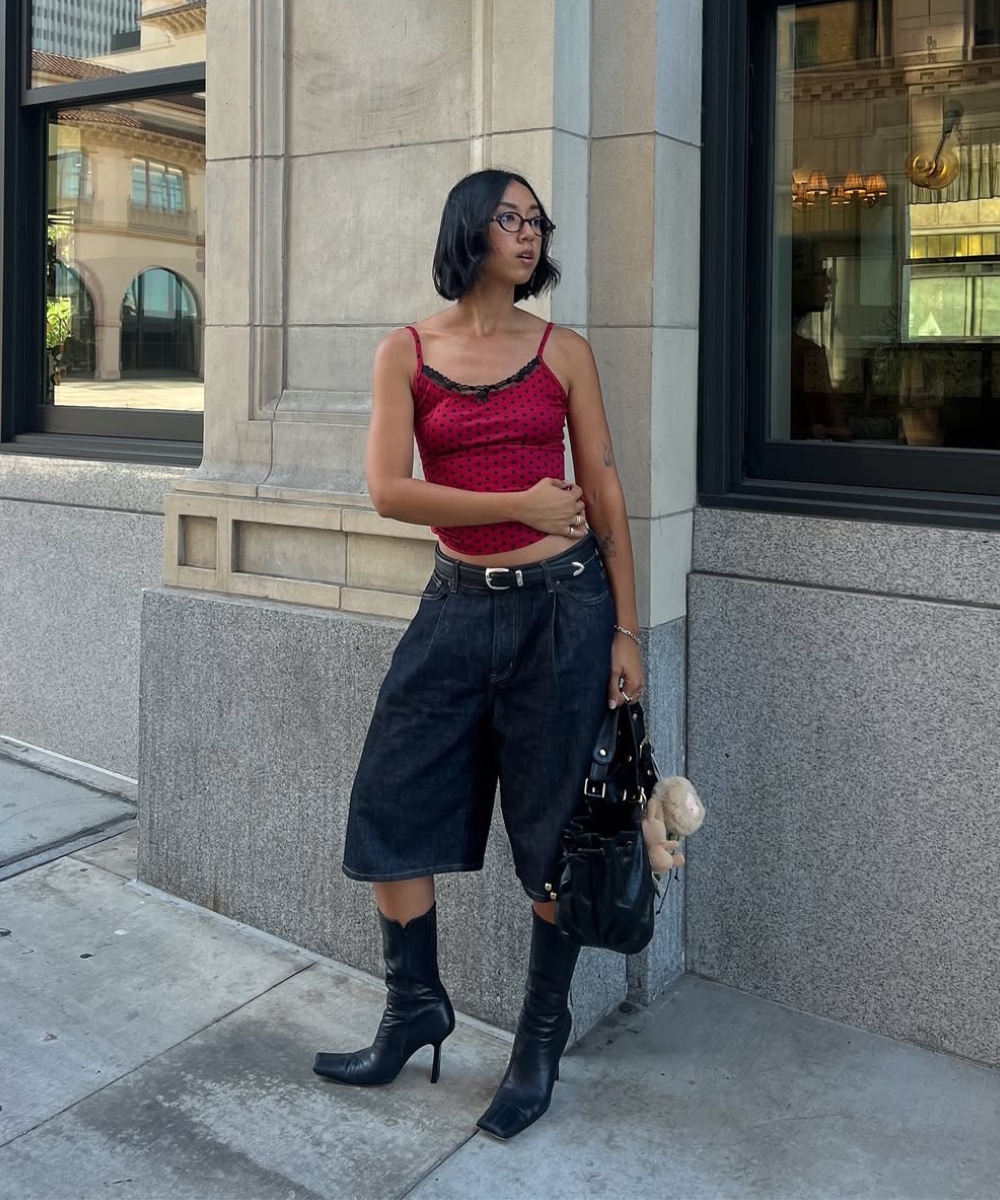Pessoa vestindo blusa vermelha e short preto com botas de salto pretas. Bolsa com chaveiro em forma de ursinho.