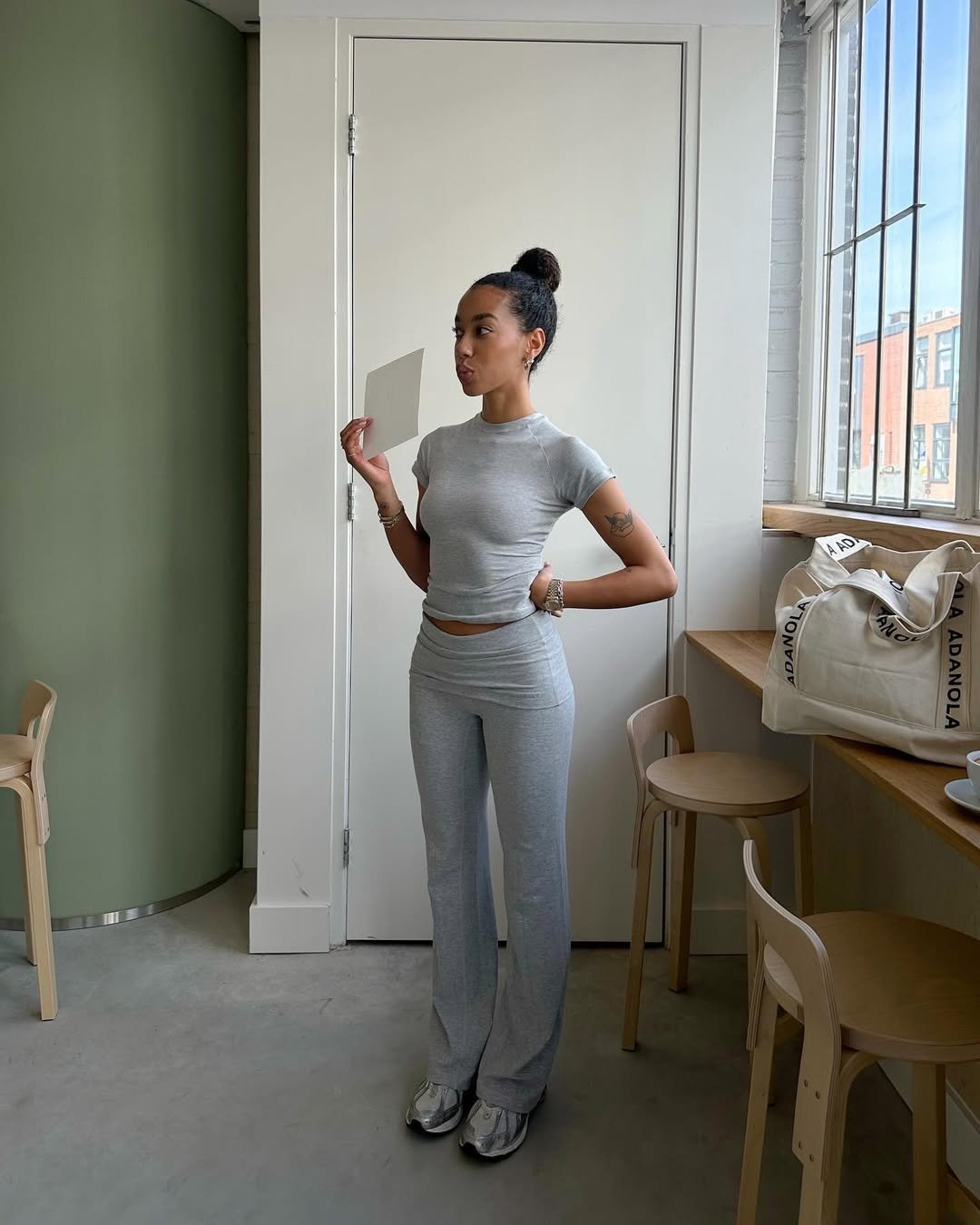 Pessoa de blusa cinza segura papel em ambiente claro. Veste calça bailarina e tênis. Bolsa na mesa ao lado.