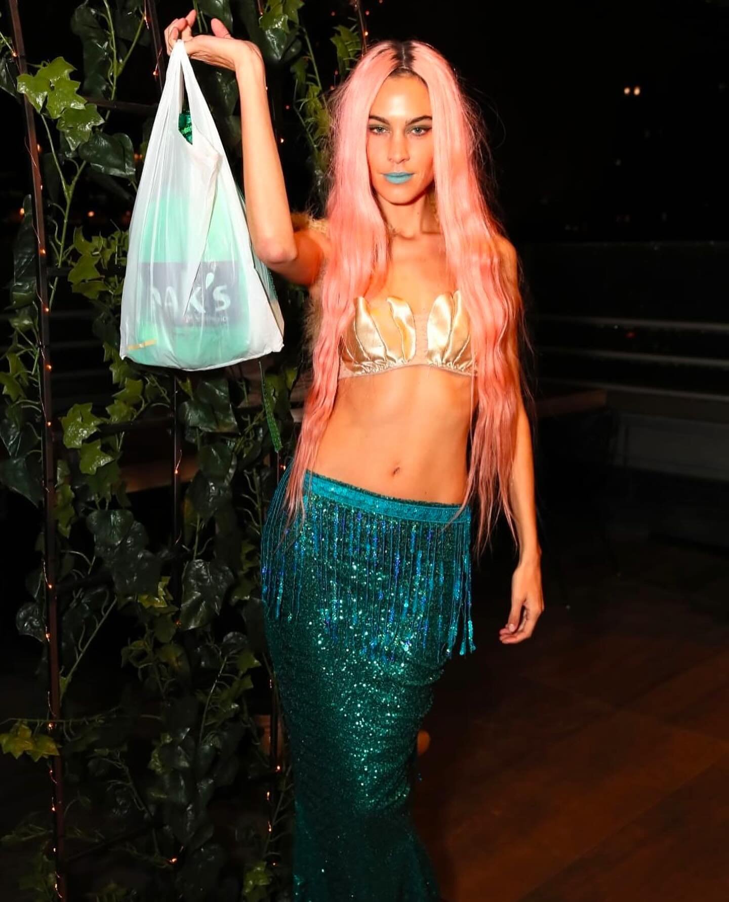 Mulher com fantasia de sereia com cabelo rosa e saia verde brilhante. Ela segura uma sacola plástica.