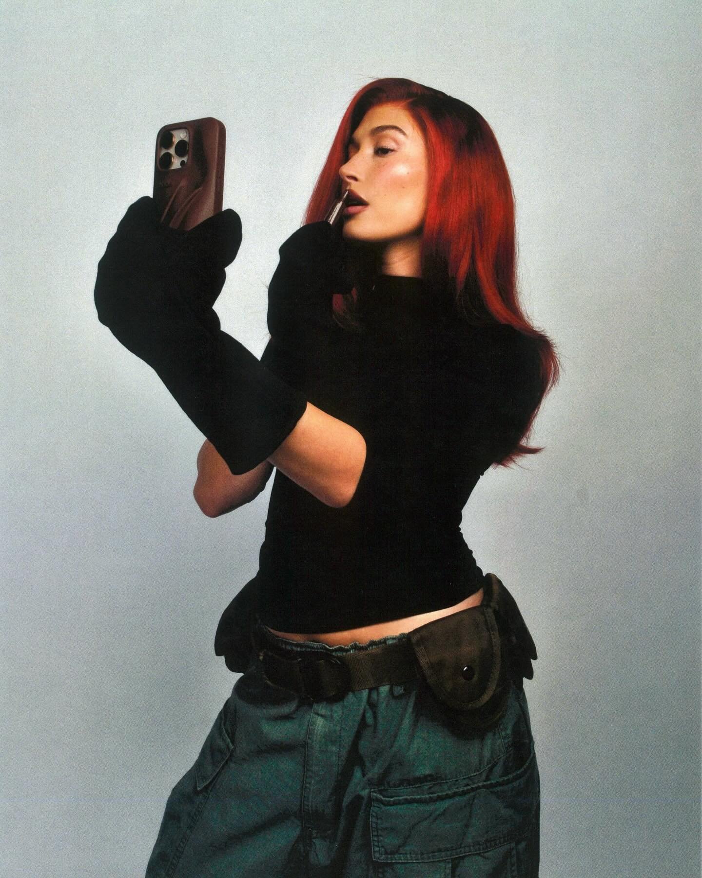 Mulher com cabelo vermelho vibrante aplica batom, segurando um smartphone, evocando fantasia de Halloween feminina.