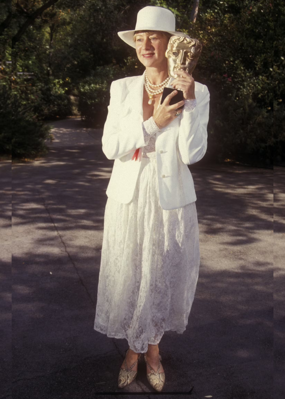 Helen Mirren segura um troféu enquanto veste um chapéu e traje branco, em ambiente externo.
