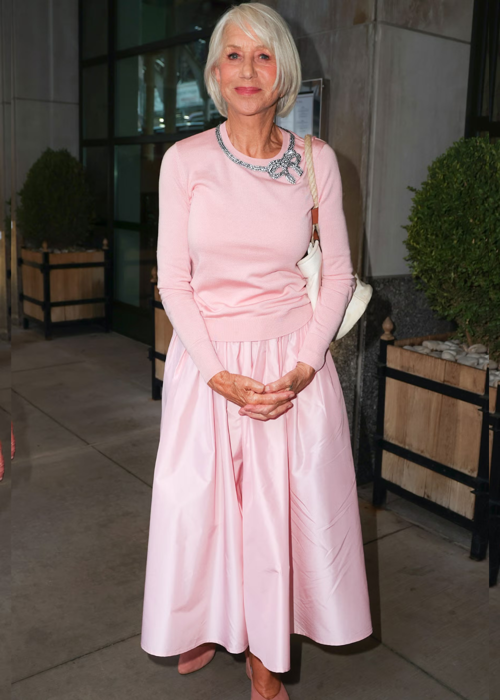Helen Mirren veste um conjunto rosa elegante com uma saia longa. Ela está em pé diante de um edifício.