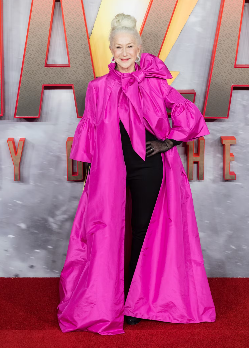 Uma pessoa em pé sobre um tapete vermelho, usando um vestido longo rosa vibrante com laço grande no pescoço.