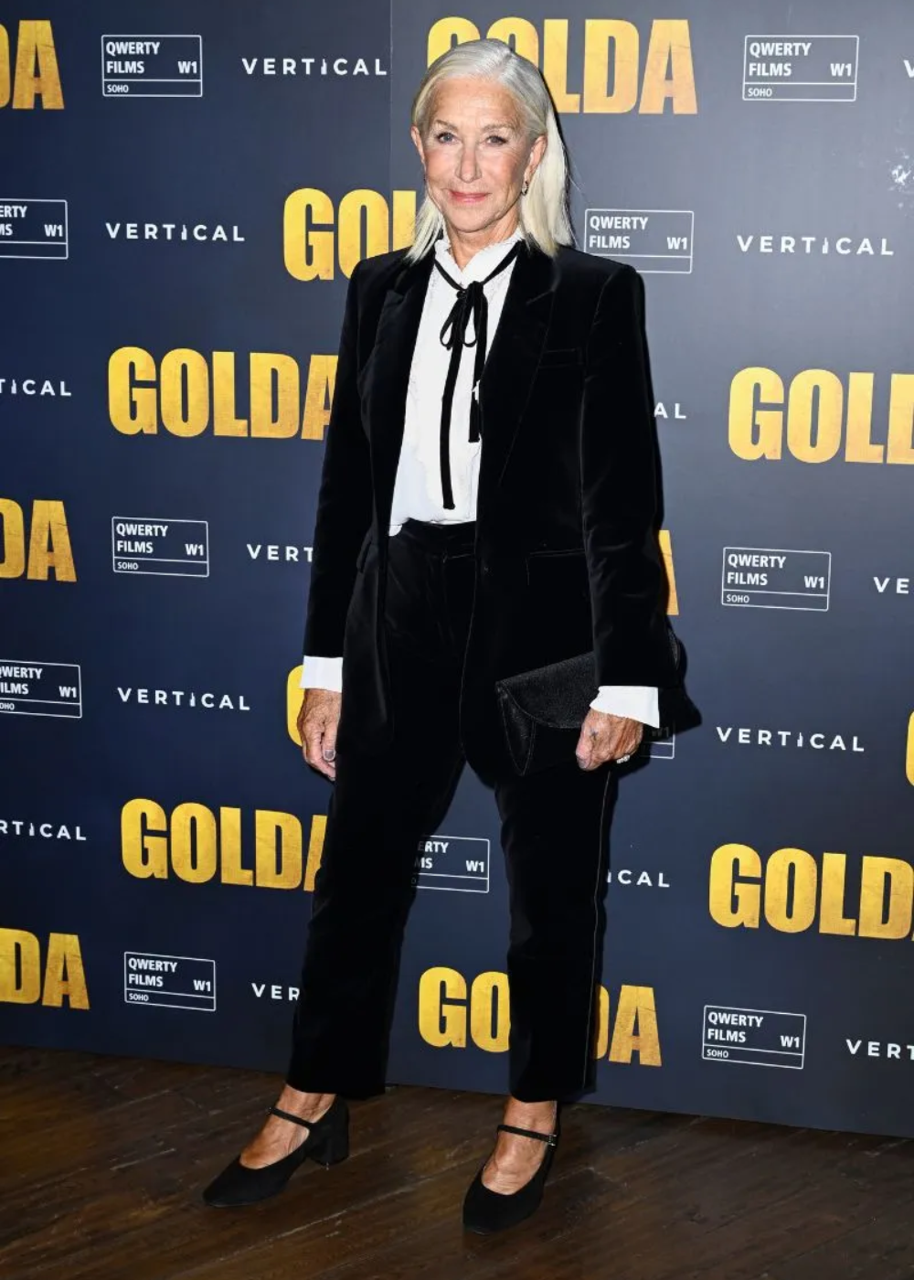 Helen Mirren de pé em frente a um painel azul com escritas em amarelo usando um terno preto com camisa branca.