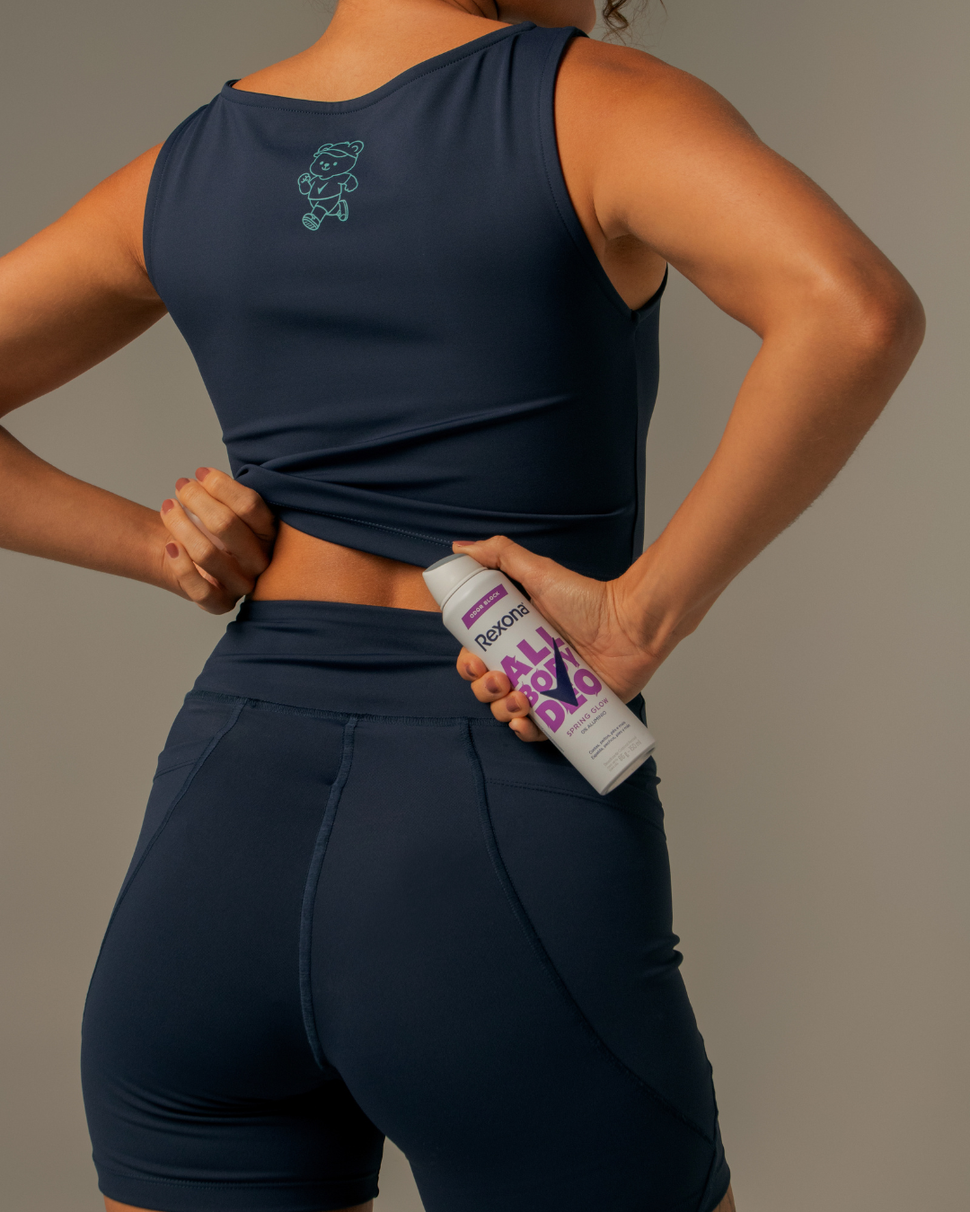 Mulher em roupa de ginástica azul segurando um desodorante corporal próximo às costas.