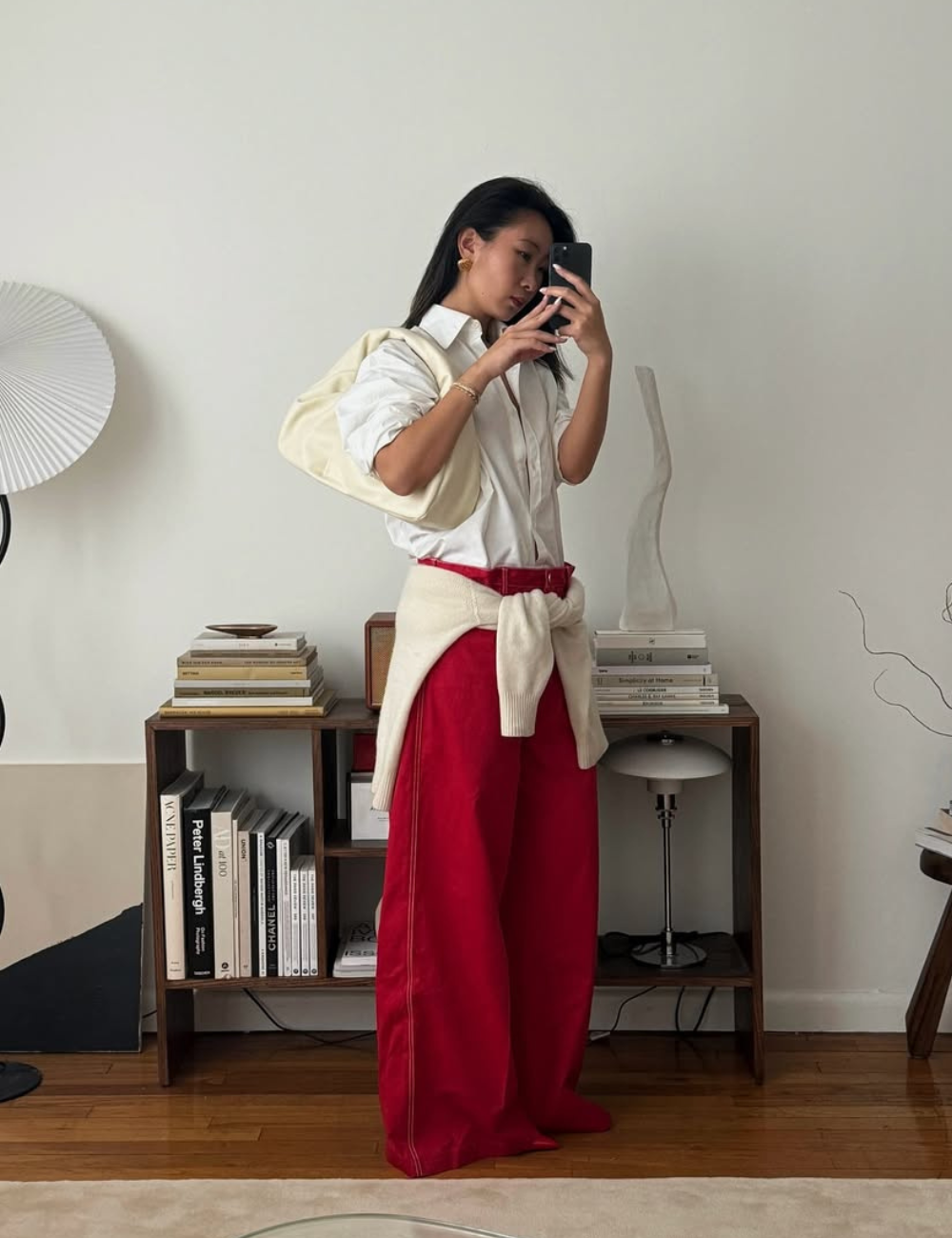 A imagem apresenta uma mulher com calça pantalona vermelha, combinada com uma camisa branca de botões. Ela usa um suéter claro amarrado na cintura, criando um visual moderno e descontraído. O ambiente é minimalista, com móveis de madeira e livros.