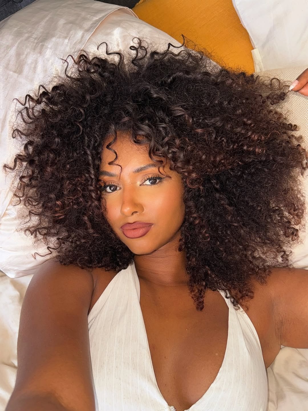 Cabelo volumoso e bem definido é o destaque da imagem, com cachos soltos e vibrantes. A modelo apresenta um tom de pele radiante, usando uma blusa clara que contrasta com o fundo neutro. Como dar volume no cabelo