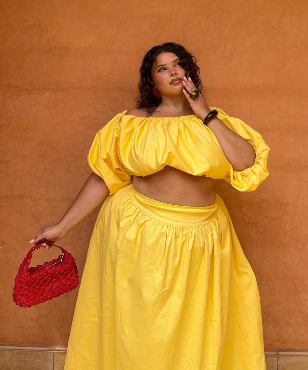 A imagem apresenta uma mulher com um look vibrante e estiloso, usando um conjunto amarelo composto por uma blusa cropped com mangas bufantes e uma saia longa, ideal para o calor. como fazer a maquiagem durar no calor