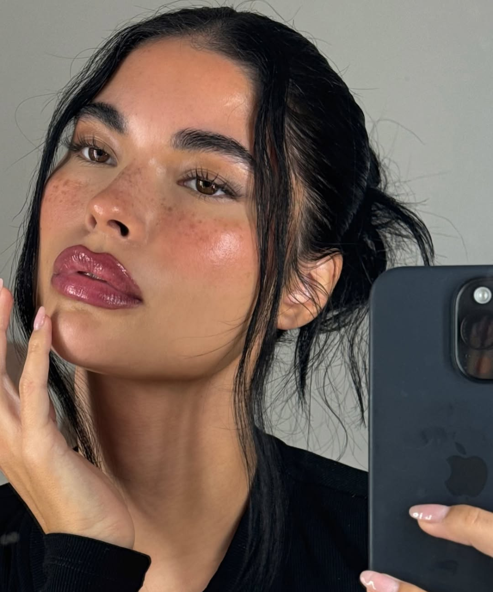 A imagem apresenta uma mulher com maquiagem leve e fresca, destacando a pele iluminada e lábios com um tom de gloss. Ela segura um celular, sugerindo um momento de selfie.