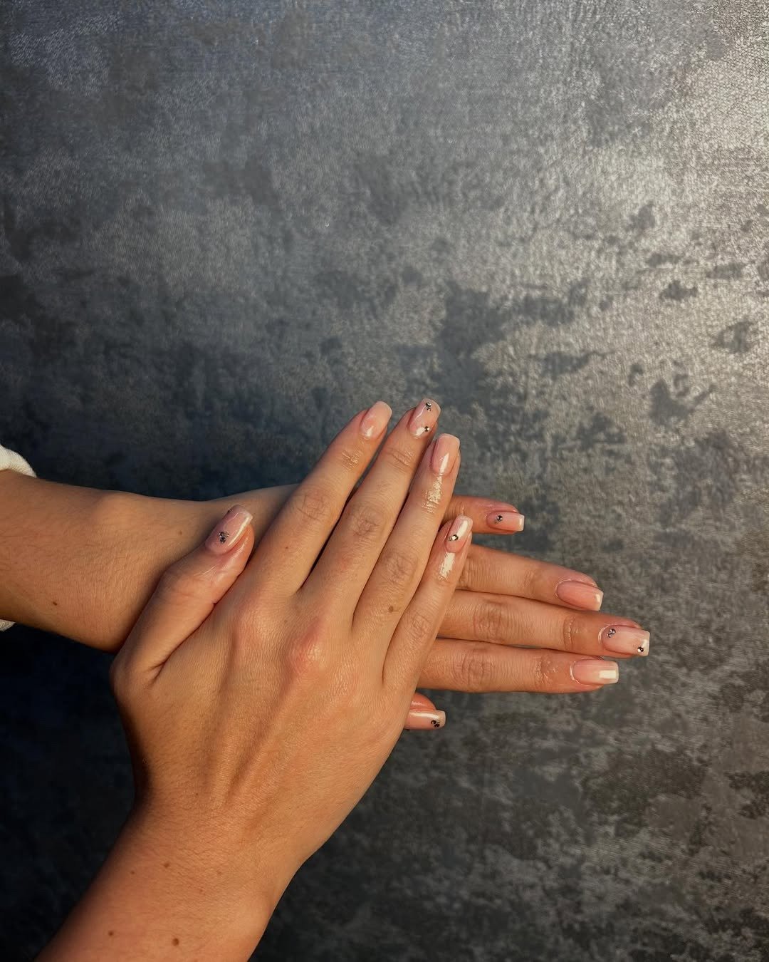 As unhas apresentam um esmalte em tom nude com detalhes em marrom e pequenas pedras decorativas, refletindo um estilo sofisticado e moderno.