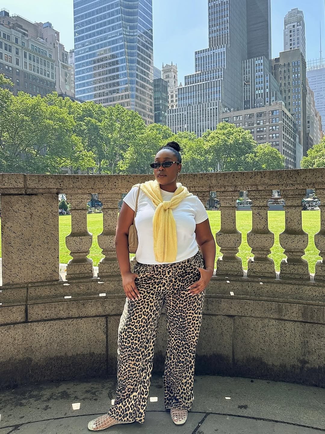 A imagem apresenta uma mulher em um ambiente urbano, com arranha-céus ao fundo. Ela usa uma camiseta branca e calças largas com estampa de onça, que trazem um toque de ousadia ao look. Um suéter amarelo está amarrado ao redor do pescoço, complementando o
