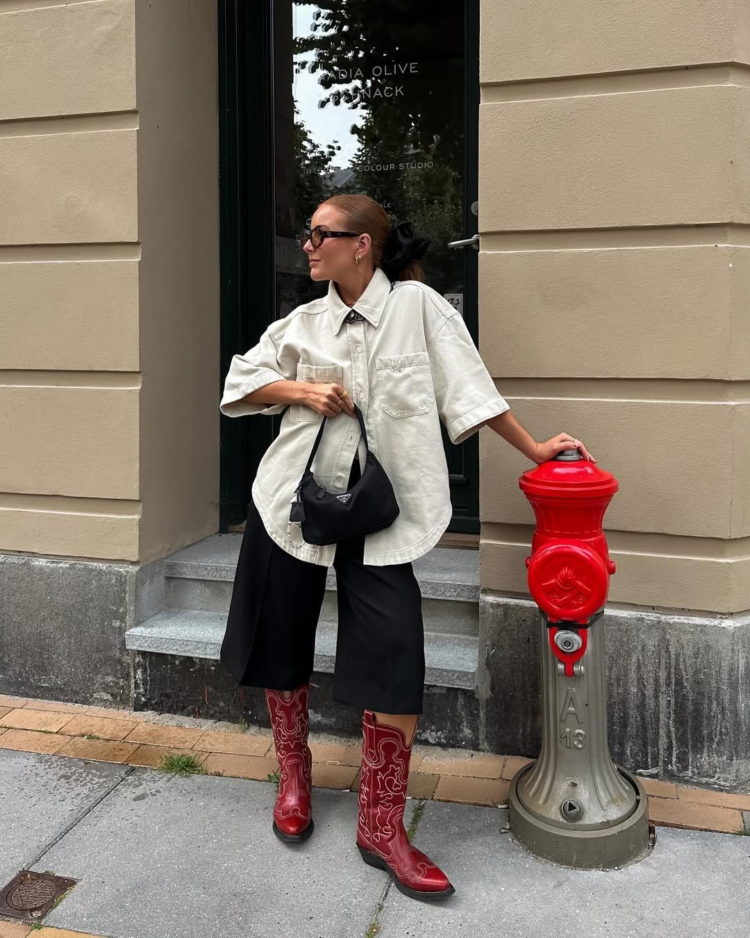 Pessoa elegante de óculos e camisa larga, usa botas vermelhas decoradas ao lado de hidrante.