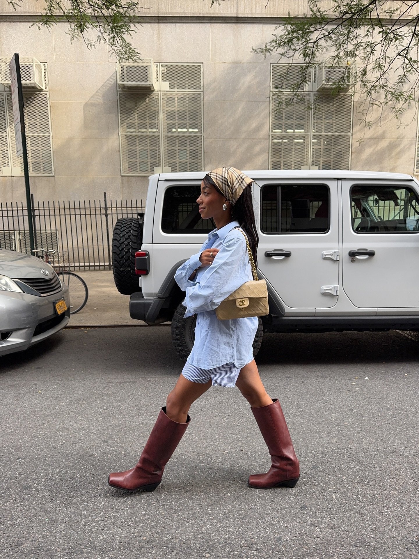Pessoa andando na rua usando botas marrons e camisa larga, com bolsa bege e lenço na cabeça.