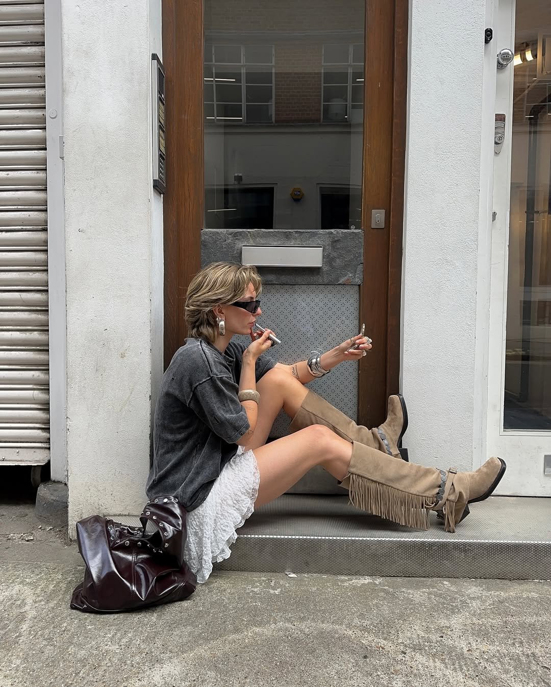 Pessoa usando botas com franjas em dia quente, sentada na calçada, retocando maquiagem.