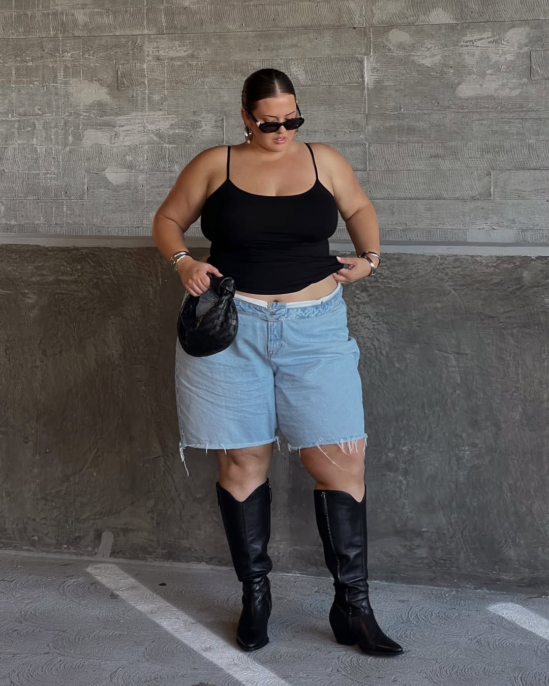 Pessoa com blusa preta, shorts jeans e bota no calor. Ela segura uma bolsa pequena em um ambiente de concreto.