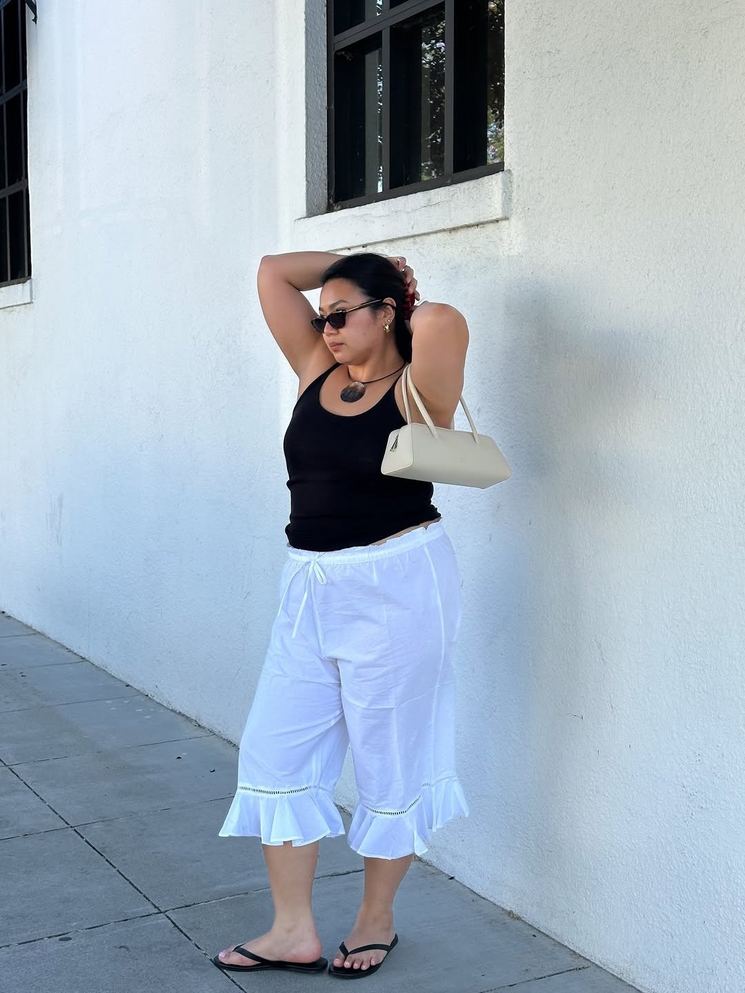 A imagem apresenta uma mulher em um ambiente urbano, vestindo uma blusa preta e calças brancas com babados na barra. Como usar chinelo