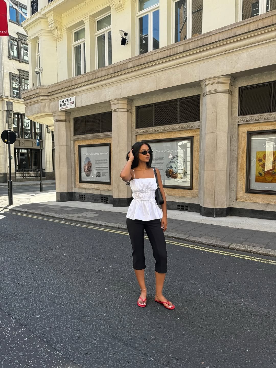 A imagem apresenta uma mulher em uma rua urbana, usando um top branco e calças pretas cropped. Ela complementa o visual com chinelos vermelhos, que trazem um toque casual e moderno ao look. Como usar chinelo