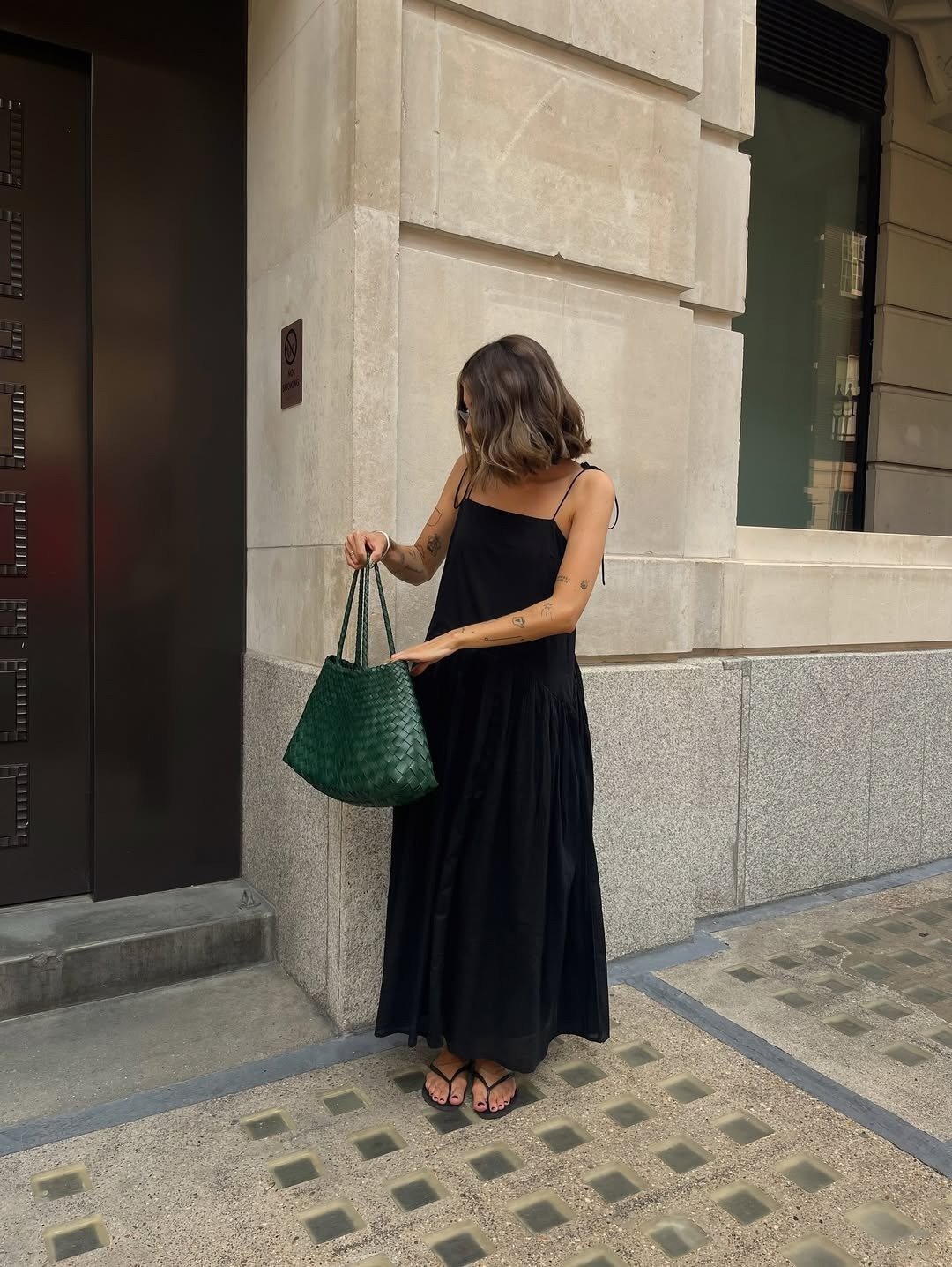 Uma mulher com cabelo ondulado e tatuagens usa um vestido longo preto, combinando com chinelos simples. Ela segura uma bolsa verde, destacando um look casual e elegante. A imagem exemplifica como usar chinelo com estilo.