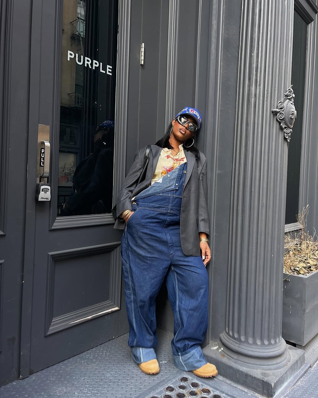 Pessoa vestindo jardineira jeans larga e jaqueta listrada, encostada em parede com portas cinzas.