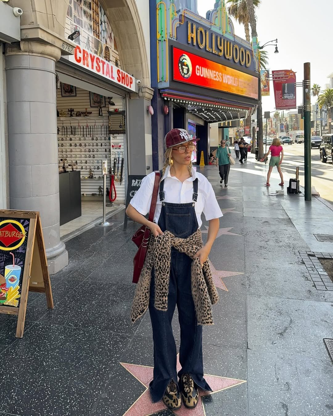 Pessoa usando jardineira jeans, camisa branca e boné, caminhar em calçada de Hollywood em frente a lojas e cinema.