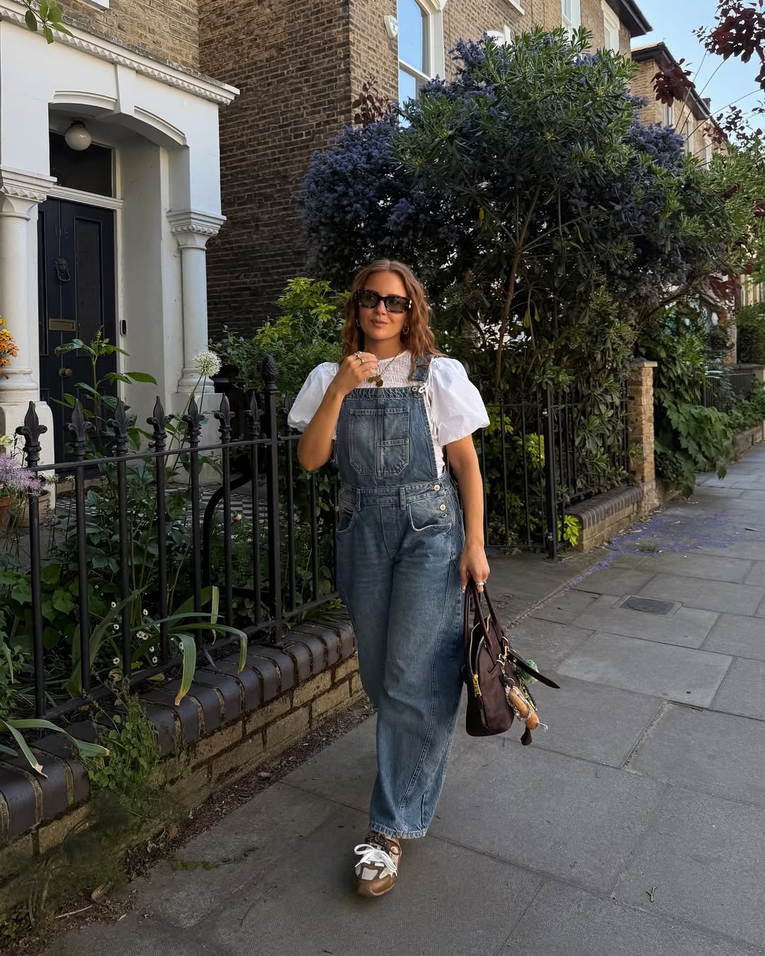 Pessoa vestindo jardineira jeans e camiseta branca, passeia em rua arborizada com casas de tijolos aparentes.