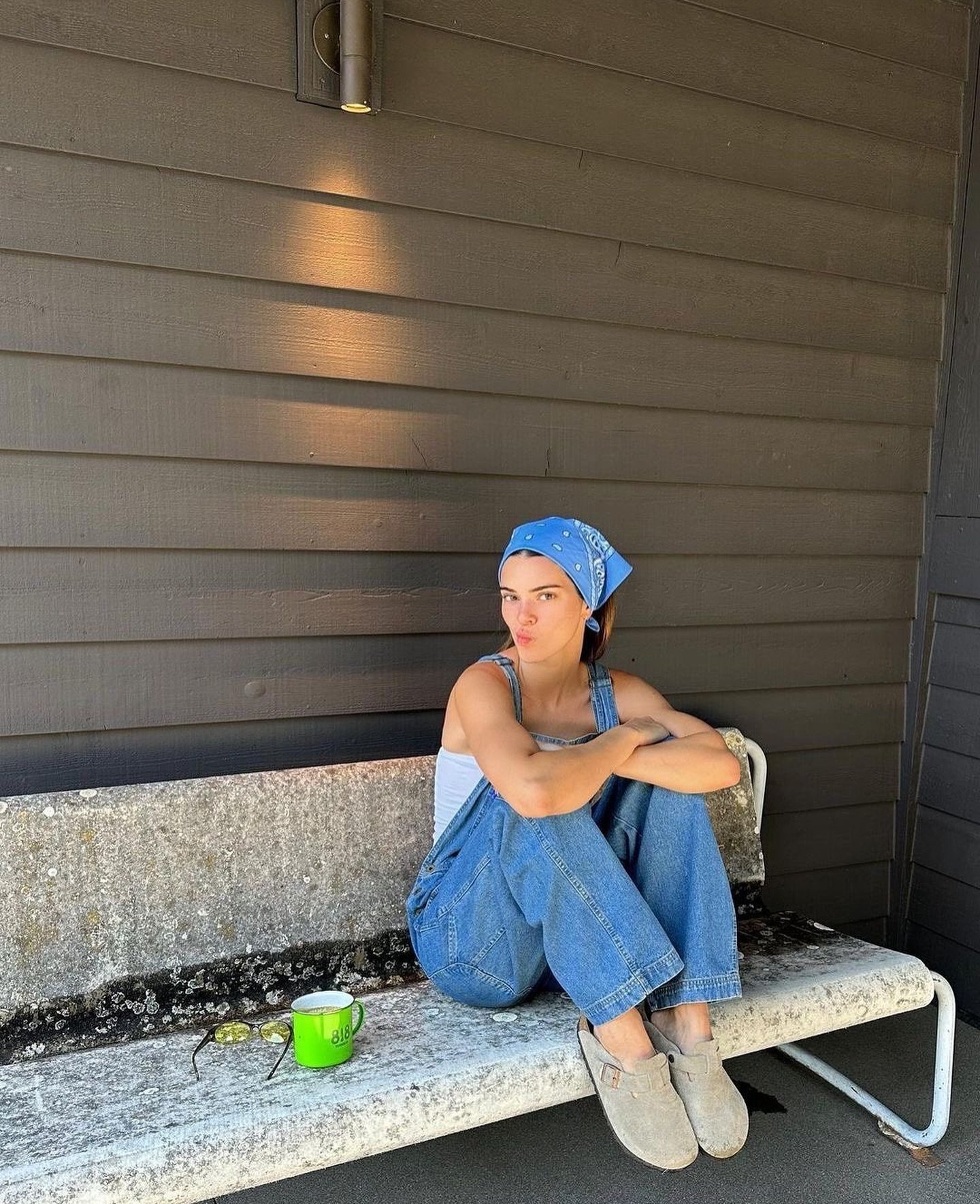 Pessoa com jardineira jeans azul e lenço sentado em banco de pedra. Há uma xícara e óculos ao lado.