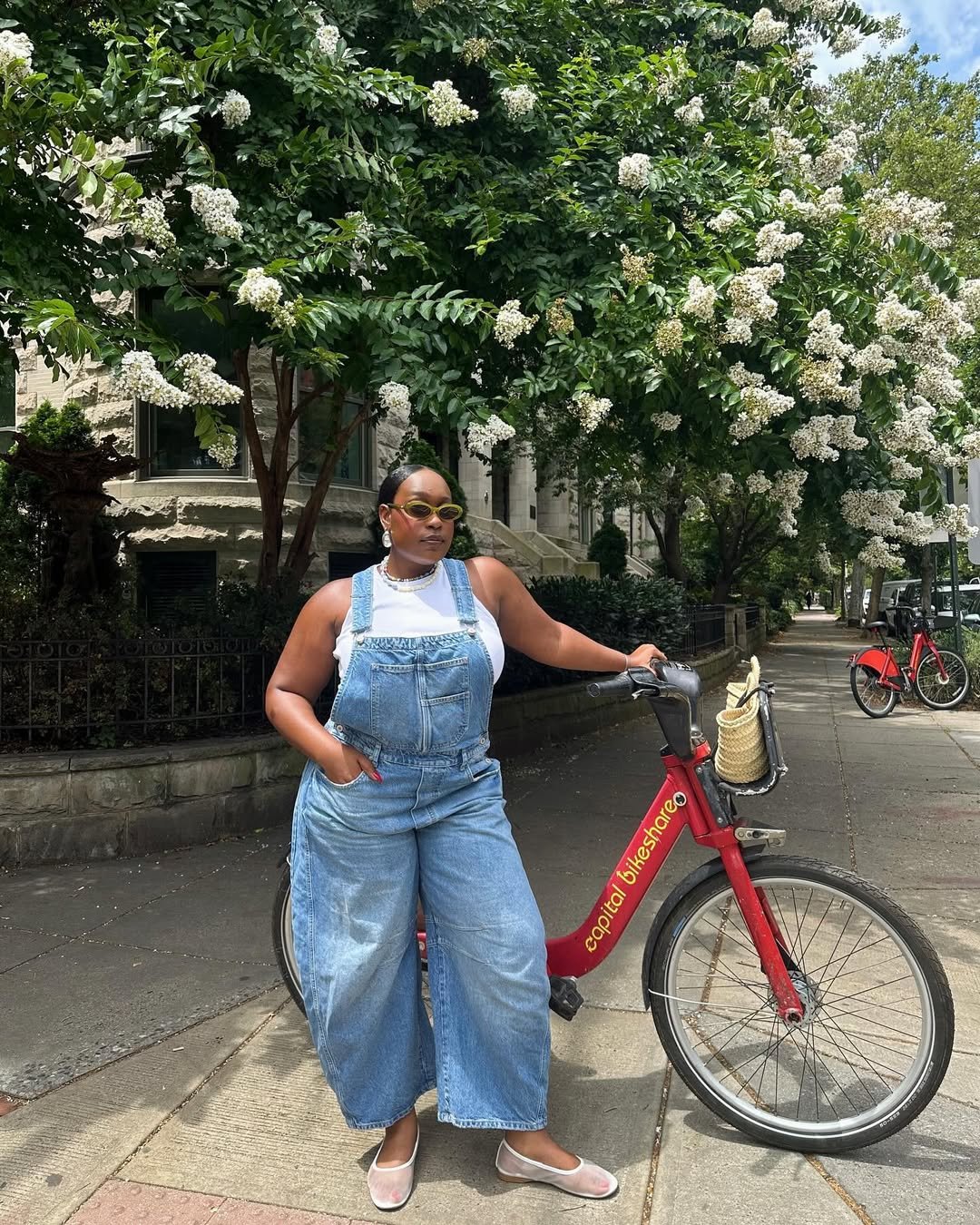 Pessoa em uma jardineira jeans posa ao lado de uma bicicleta vermelha em rua arborizada.