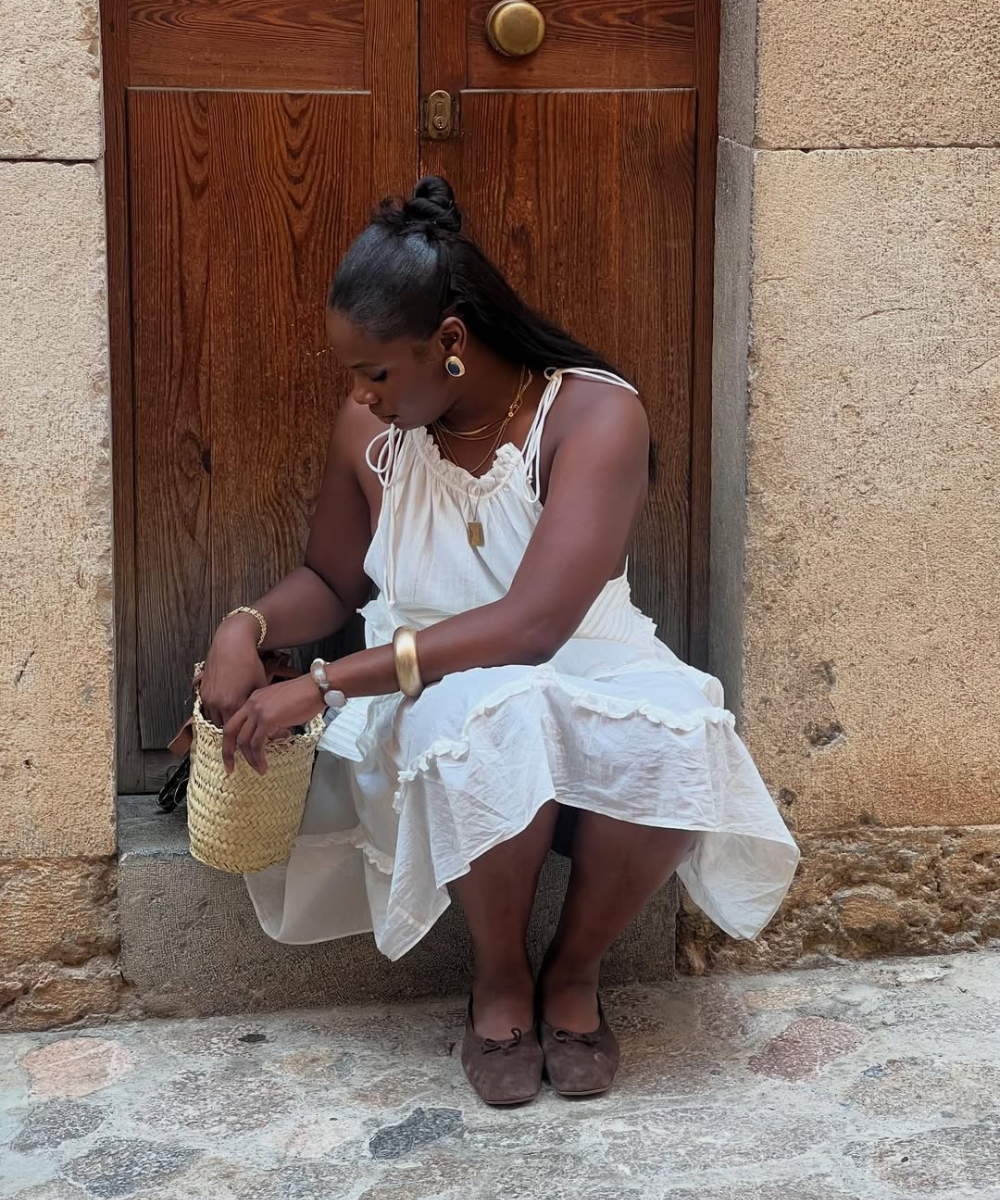 Mulher sentada em frente a uma porta de madeira, usando vestido branco, bolsa de palha e sapatilhas marrons.