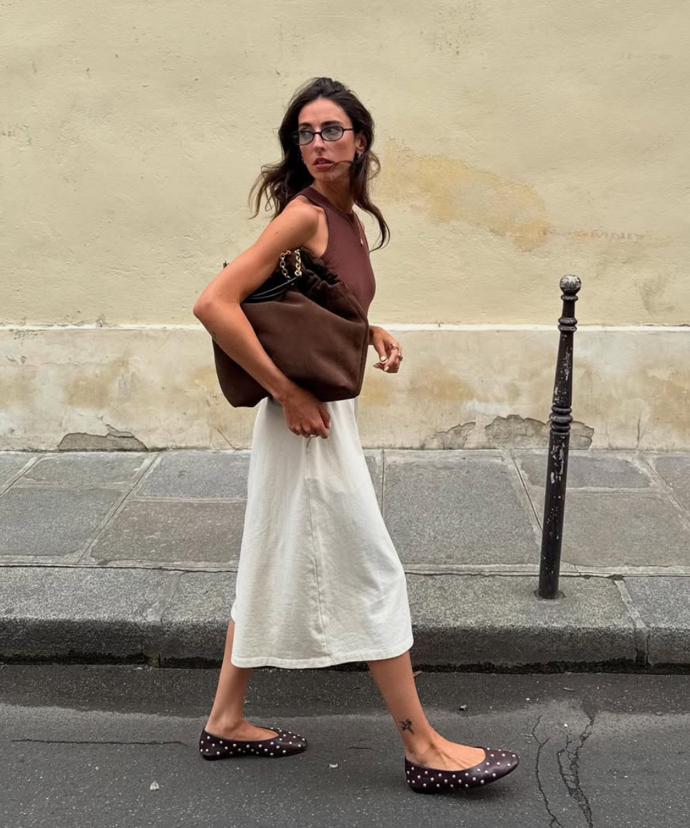 Pessoa caminha usando bolsa e sapatilhas marrons de bolinhas. Veste blusa marrom e saia branca.