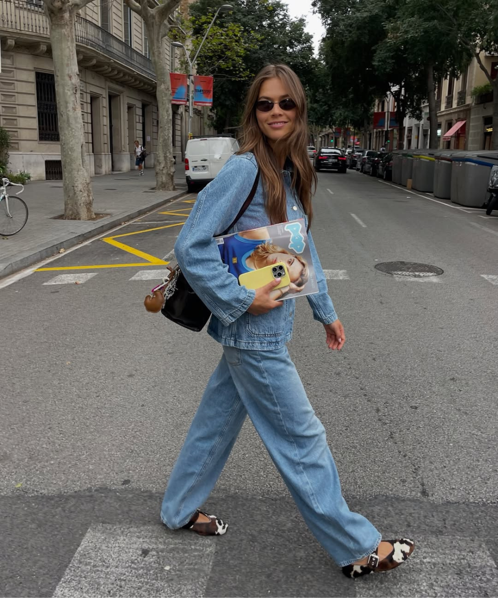 Pessoa usando conjunto jeans largo, óculos escuros e carregando uma revista. As sapatilhas marrons estão em destaque no look casual.