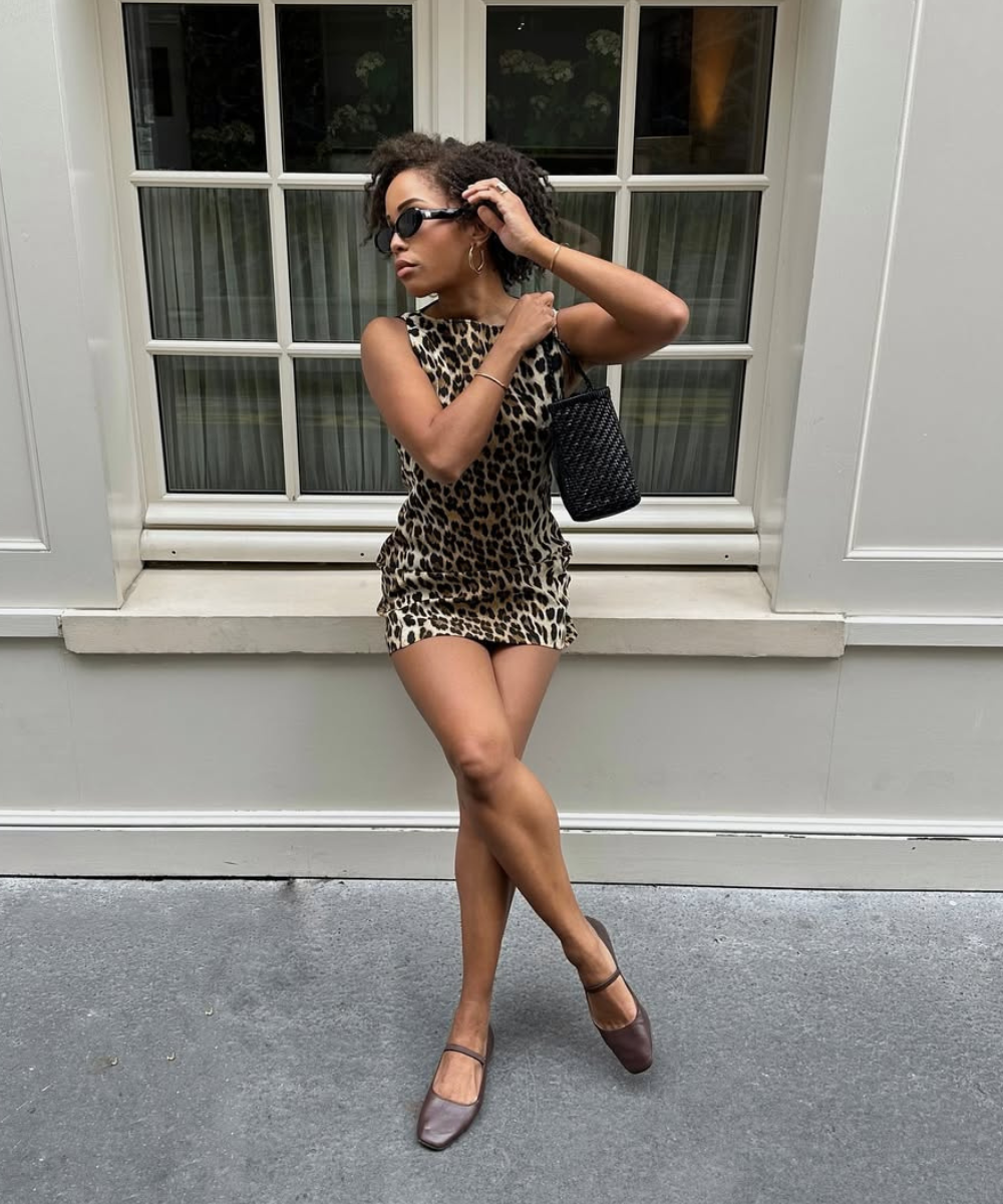Pessoa de vestido estampado segura bolsa, usa óculos escuros e sapatilha marrom, encostada em parede com janela.