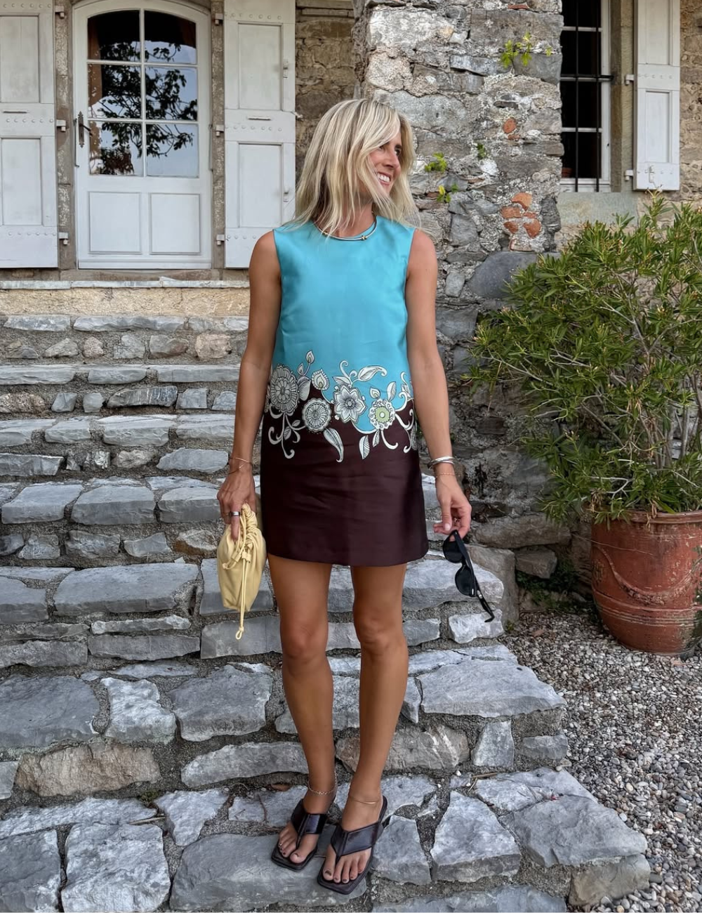 A imagem mostra uma mulher sorridente, usando um vestido azul com detalhes florais em uma tonalidade marrom, ideal para um dia ensolarado. Ela combina o look com tamancos pretos, que trazem conforto e estilo, perfeitos para passeios.