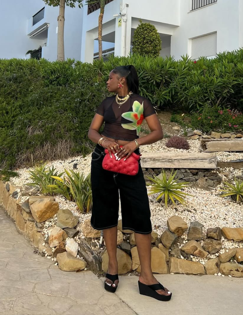 A imagem apresenta uma mulher em um ambiente ao ar livre, cercada por vegetação. Ela usa um top transparente com estampa de flor, shorts pretos e tamancos de plataforma pretos.