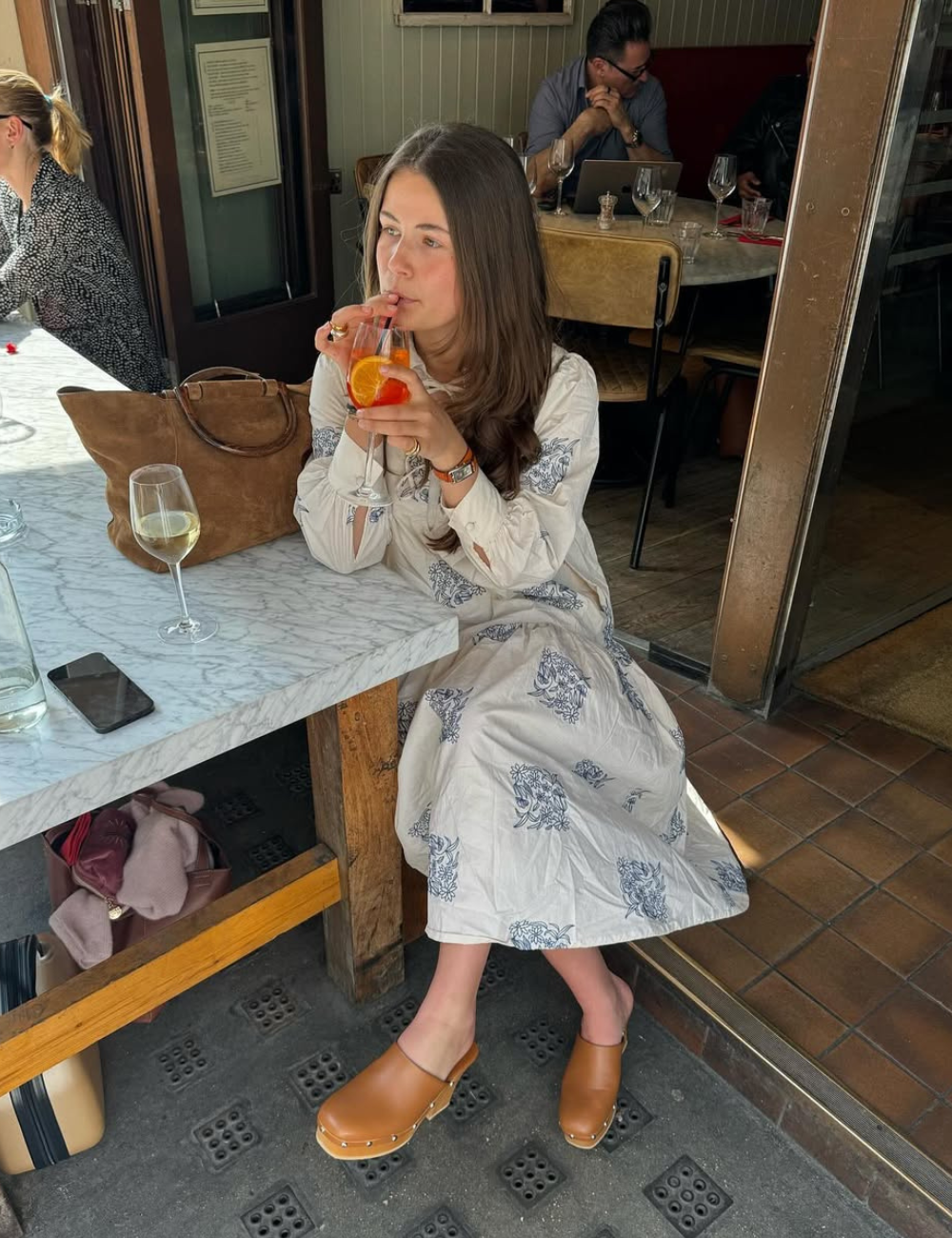 A imagem apresenta uma mulher sentada em um ambiente de restaurante, usando um vestido claro com estampas florais e tamancos de couro marrons. Ela segura um copo com uma bebida decorada com fatias de laranja, transmitindo um ar casual e elegante.