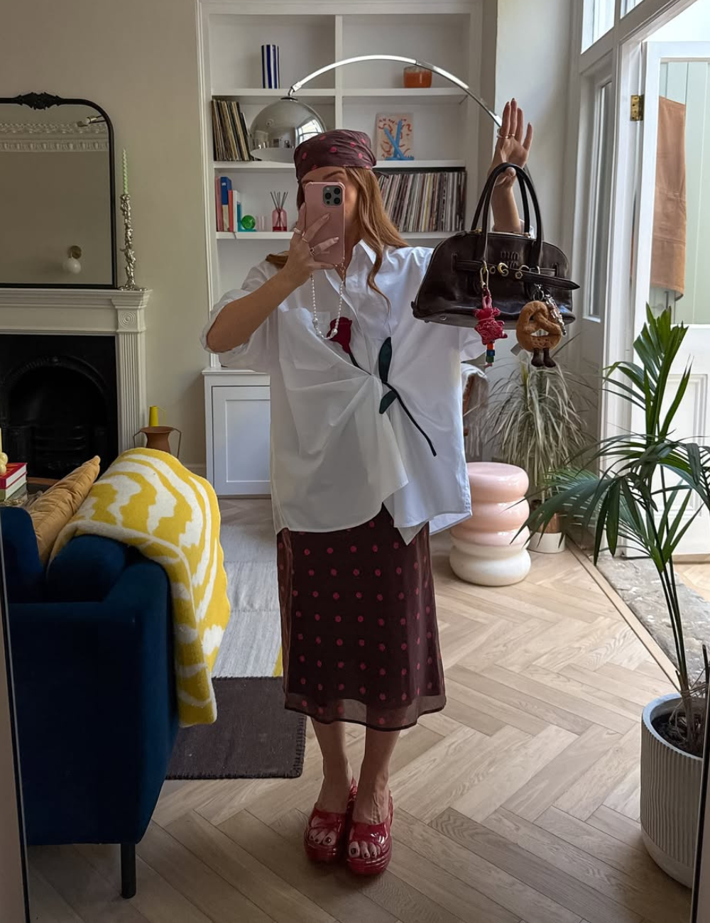 A imagem mostra uma pessoa em um ambiente interno, refletida em um espelho. Ela usa uma camisa branca oversized com um detalhe de estampa floral, uma saia marrom com bolinhas rosas e tamancos vermelhos.