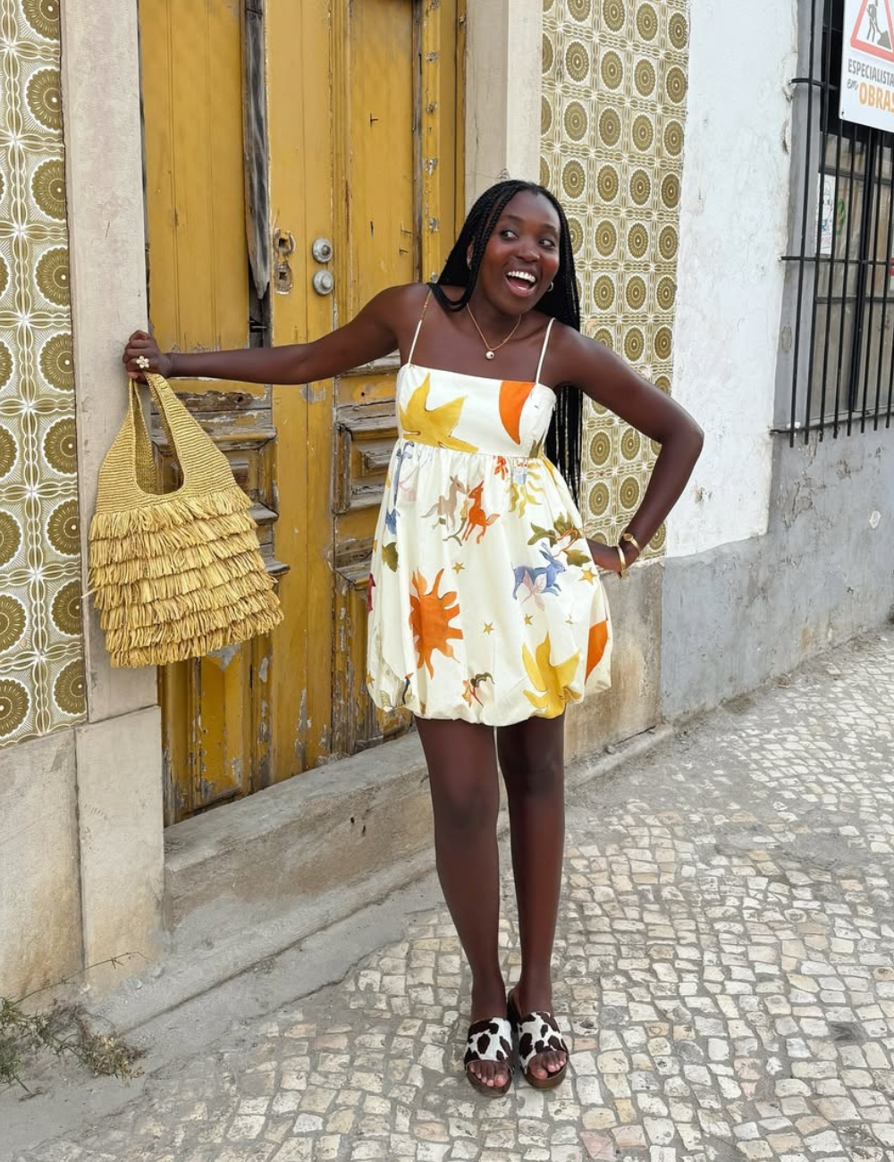 A imagem apresenta uma mulher sorridente, usando um vestido leve e colorido, ideal para dias quentes. O vestido tem estampas vibrantes e um corte solto, complementado por um tamanco estiloso com estampa de vaca.