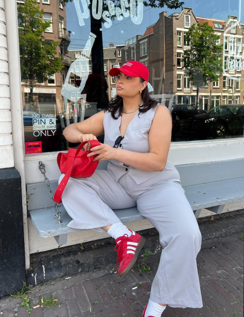 A imagem apresenta uma mulher sentada em um banco, usando um conjunto cinza e tênis vermelhos com detalhes brancos. Ela segura uma bolsa vermelha e usa um boné vermelho, criando um visual harmonioso e moderno.