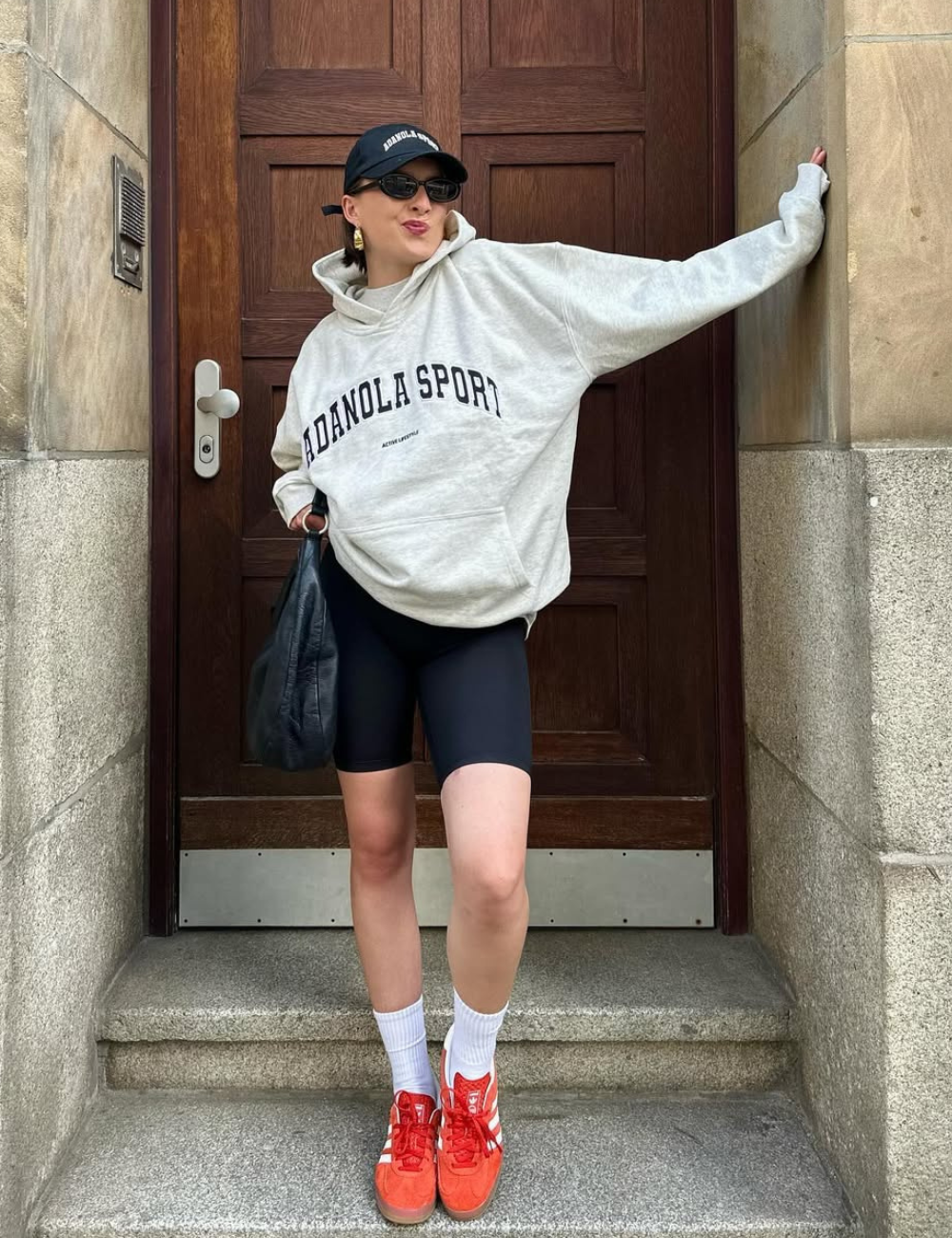 A imagem apresenta uma jovem em um ambiente urbano, usando um moletom cinza e shorts pretos. O destaque fica para os tênis vermelhos, que adicionam um toque vibrante ao look casual.