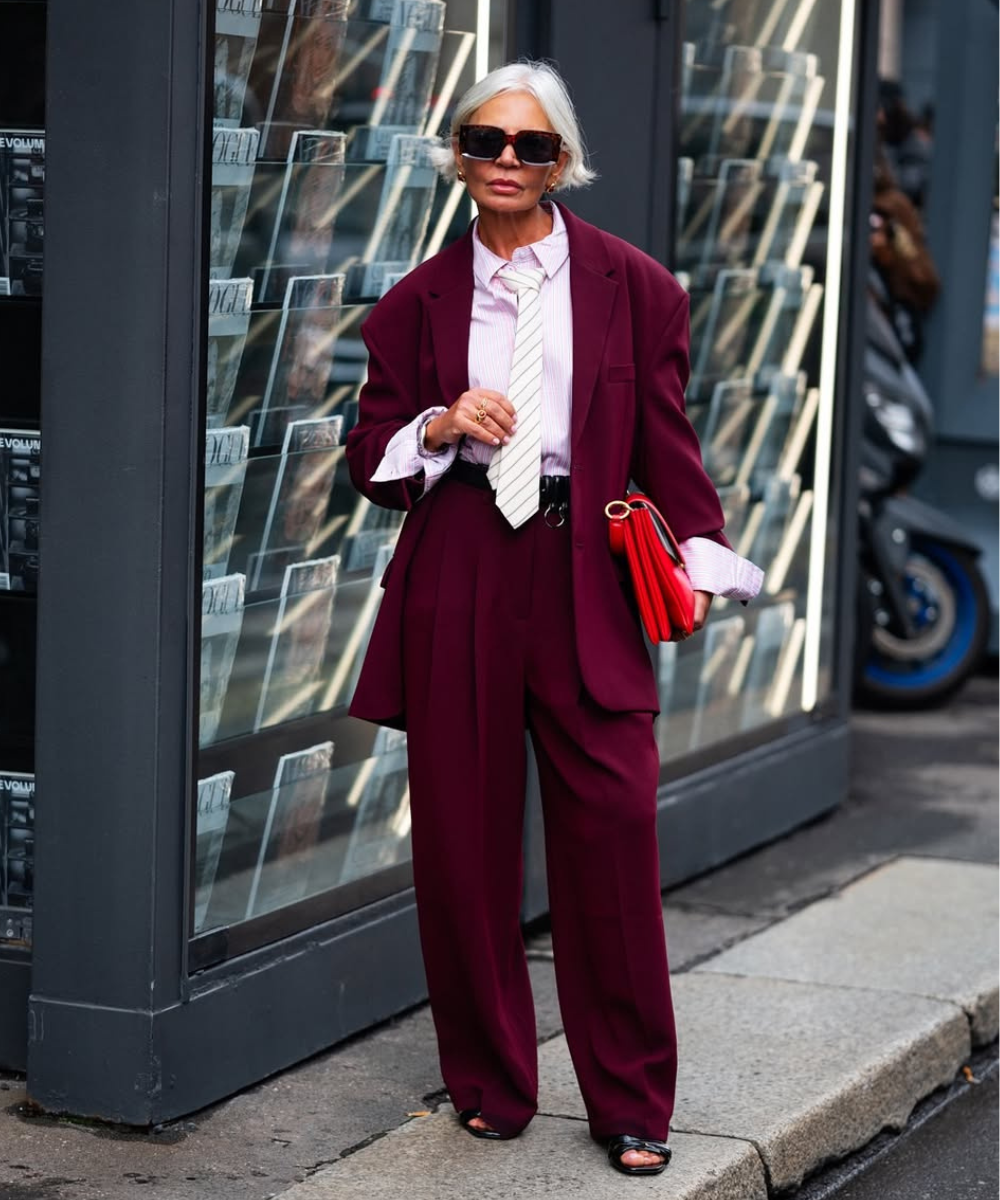 Mulher elegante com terno de alfaiataria vinho, camisa listrada rosa e gravata branca segurando uma clutch vermelha. Uma das cores de calça de alfaiataria tendência.