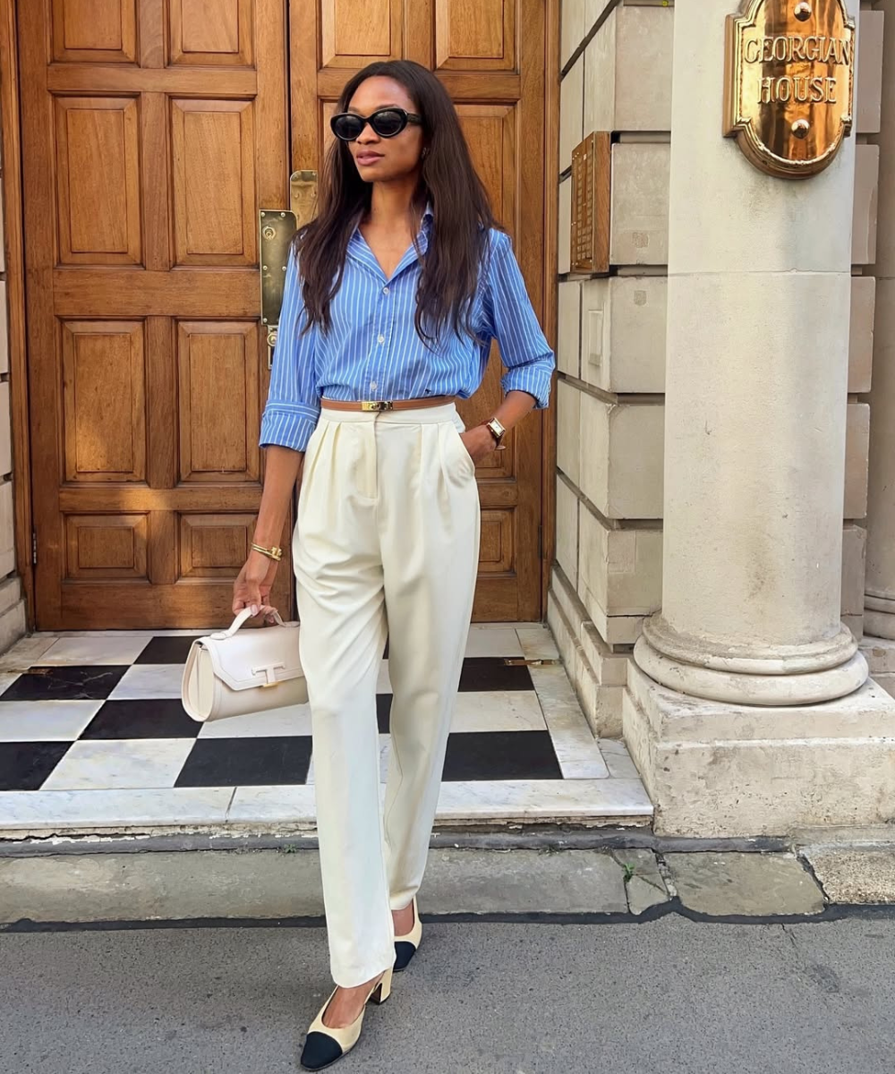 Mulher usando camisa listrada azul e calça de alfaiataria branca, segurando bolsa. Fundo com porta de madeira.