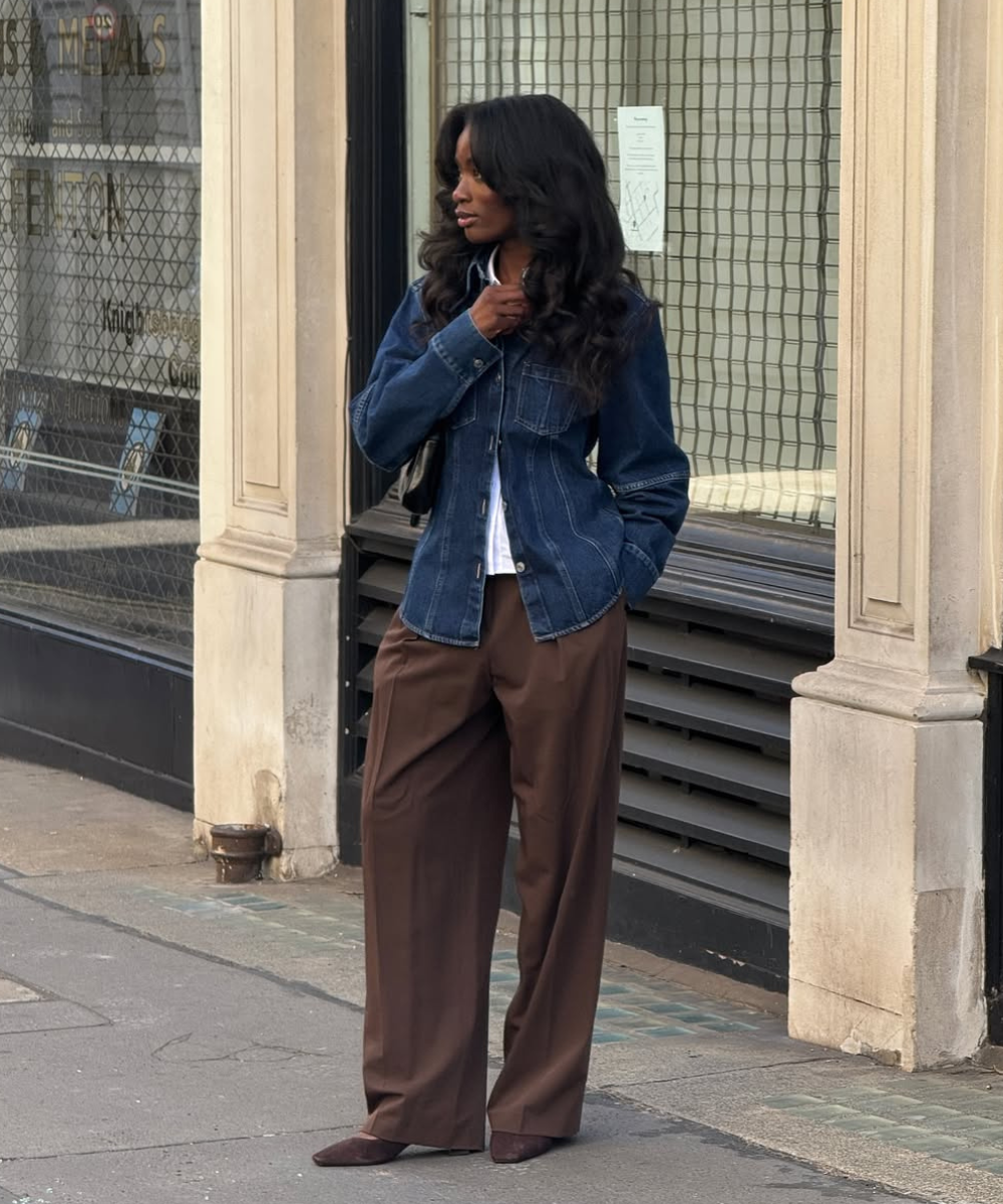 Pessoa com jaqueta jeans e calça de alfaiataria marrom, de pé em calçada urbana.