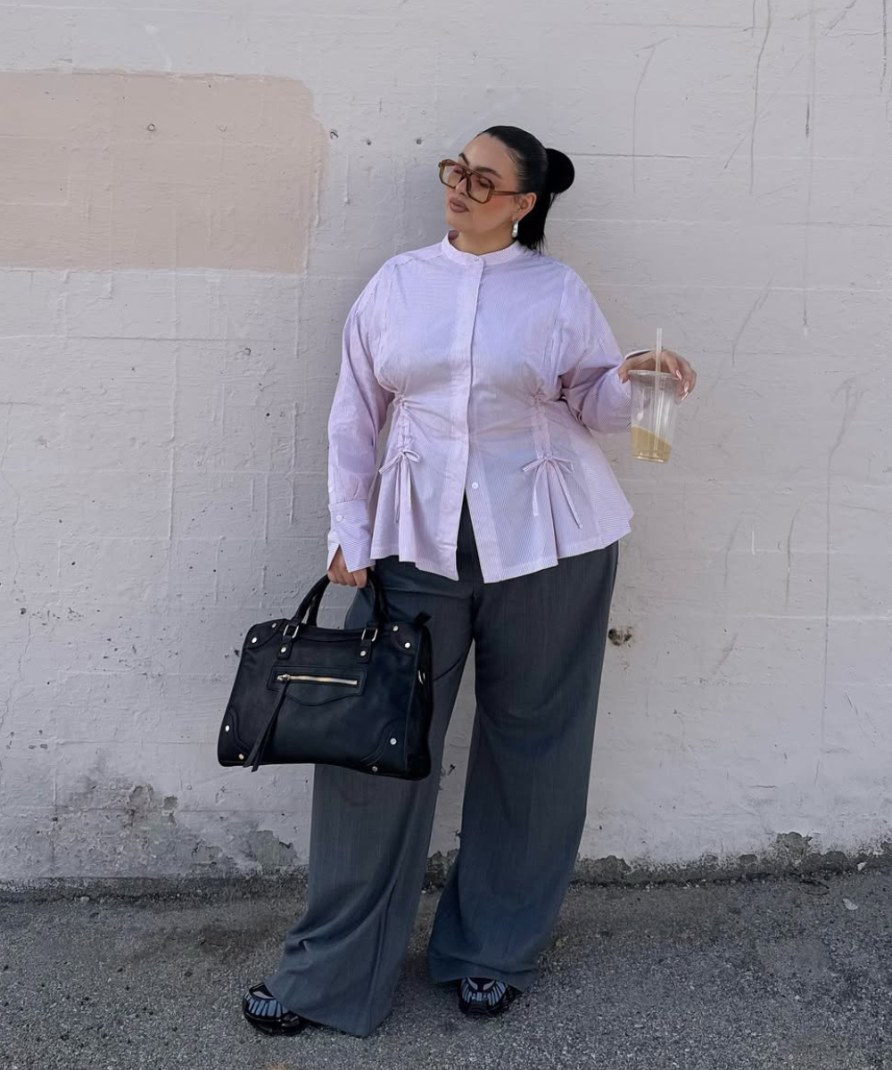 Pessoa vestindo camisa lilás e calça cinza de alfaiataria, segurando bolsa preta e bebida.