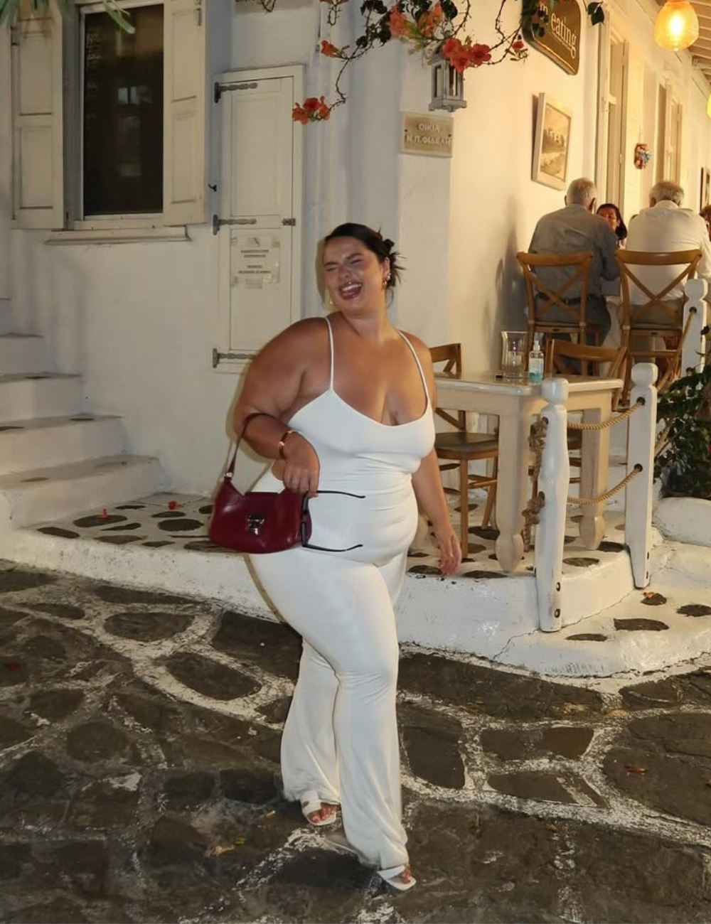 Uma mulher usa um macacão branco justo, ideal para noites de verão, combinando com rasteirinhas elegantes. Acessórios como uma bolsa vermelha adicionam um toque de cor ao look.