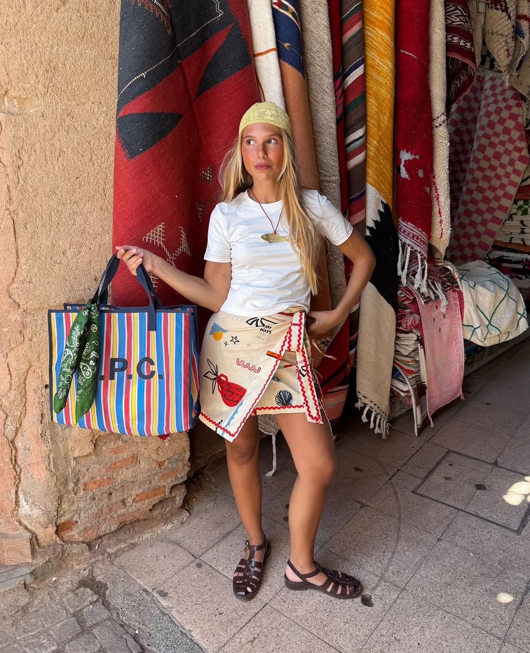Mulher com sandálias marrons segurando bolsa listrada em frente a tapetes coloridos. Simbolizando cores de sandália para o verão.
