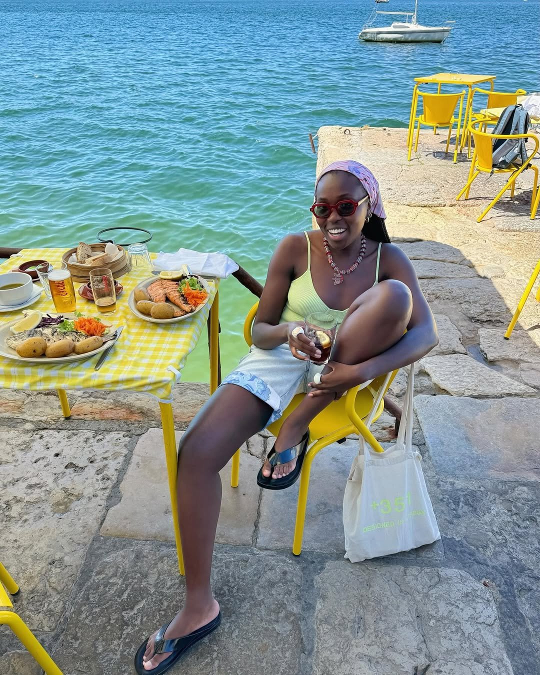 Pessoa sentada em uma cadeira amarela à beira da água, usando sandálias escuras. Mesa com comida e bebida ao lado.