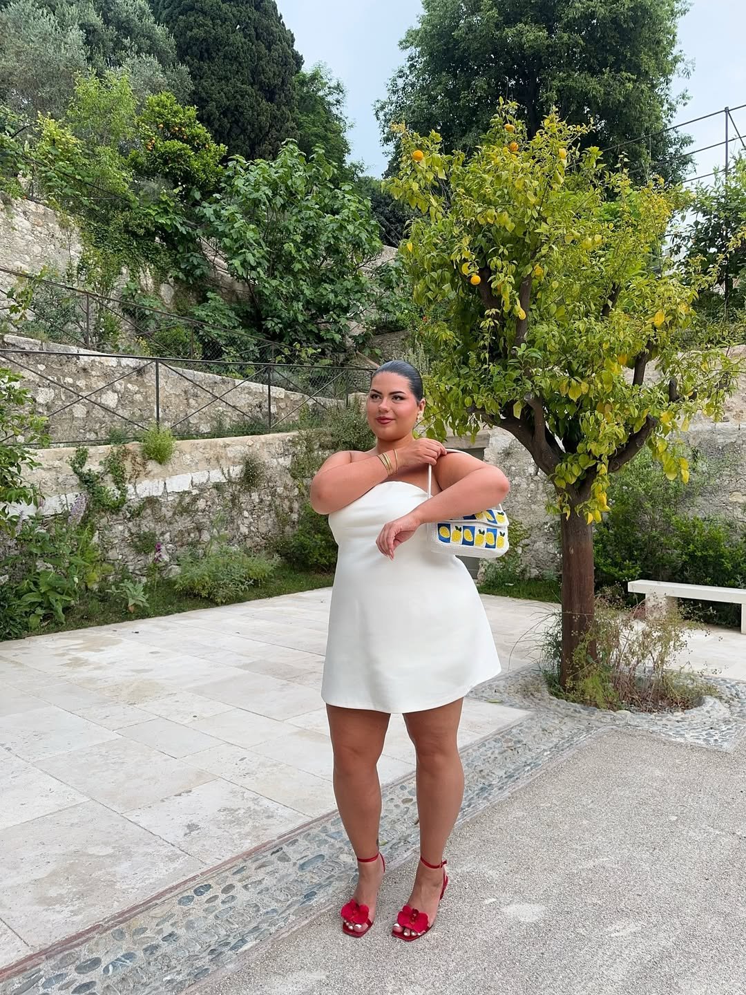 Pessoa em vestido branco e sandália vermelha, posando ao ar livre com árvores e vegetação ao fundo.