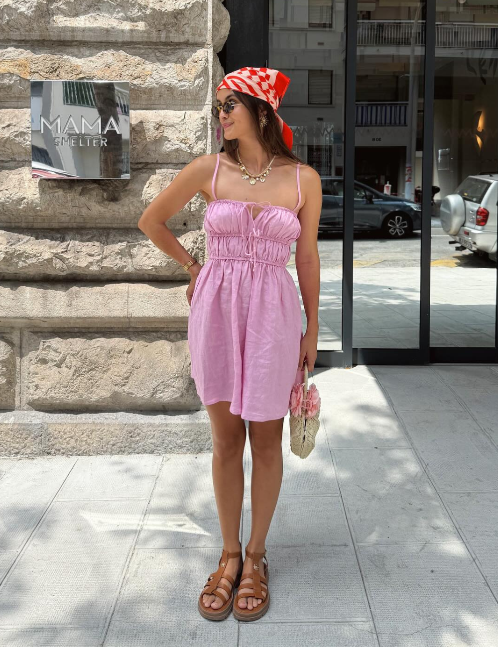 Mulher com vestido rosa claro e lenço vermelho na cabeça, em pé na frente de prédio de pedra. Cores de vestido para o verão e primavera.