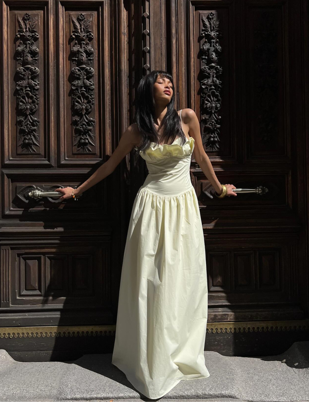 Pessoa usando vestido creme em frente a uma porta de madeira ornamentada, destacando as cores de vestido suaves sob a luz do sol.