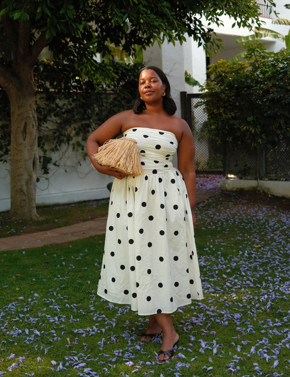 Vestido branco com bolinhas pretas, usado por uma mulher, em um jardim com flores roxas caídas.