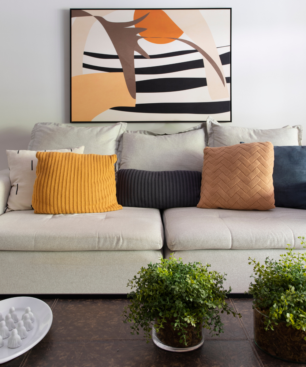 Sofá cinza com almofadas coloridas e quadro abstrato no fundo. Toques coloridos na decoração, com plantas ao redor.
